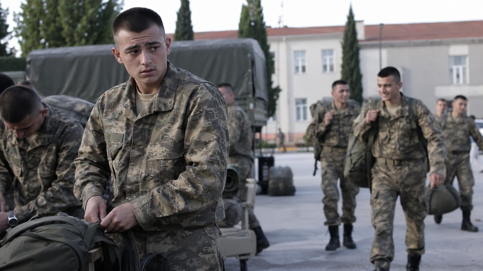 Kışla avlusunda eğitim hazırlığı yaparken yeni düzene alışmaya çalışan Türk asker adayı ve daha rahat hareket eden diğer personel
